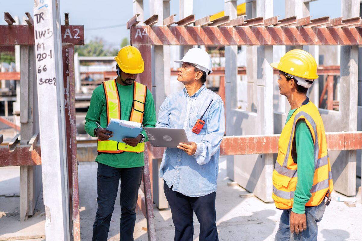 supervisor holding laptop checks quality at construction site as two workers look on