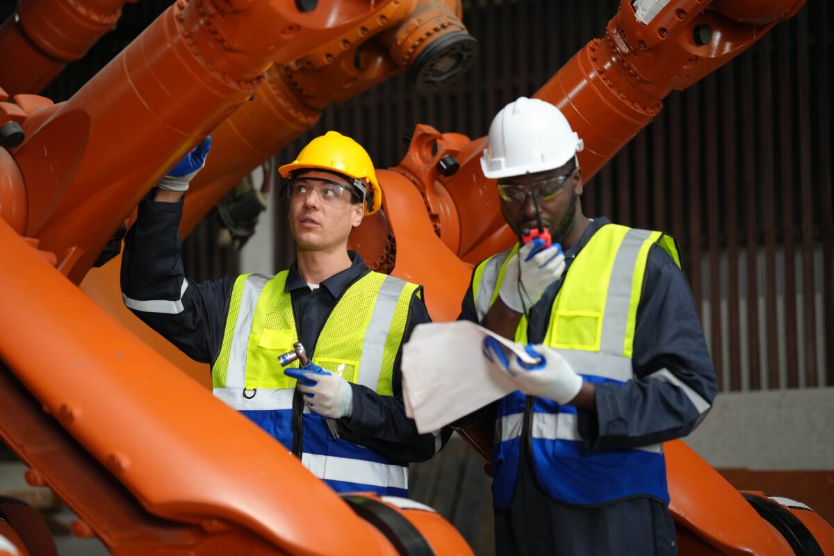 two workers standing near heavy machinery, one holding a spanner and other talking on walkie talkie
