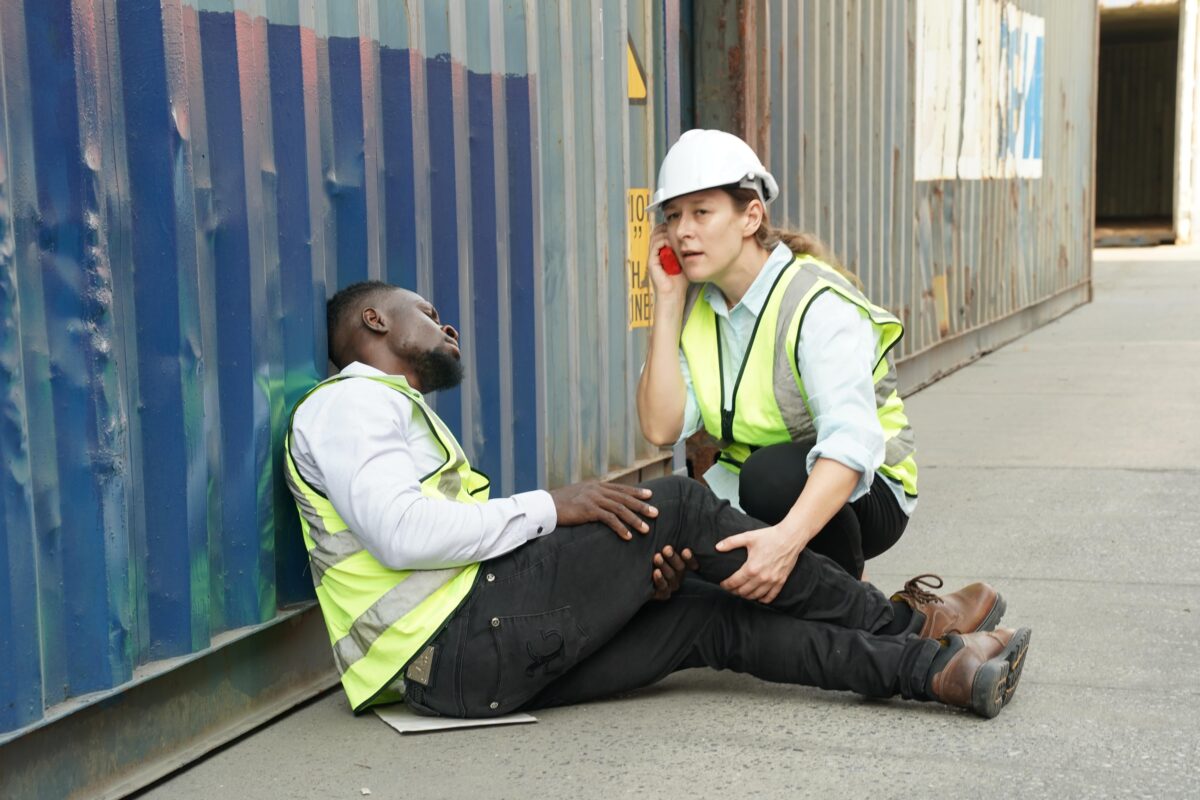 injured worker leans on cargo container as colleague calls for help